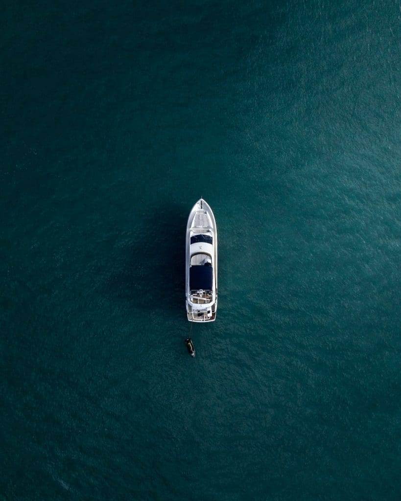 Aerial view of a white yacht anchored in deep blue-green water. A small dinghy is tethered to the side of the yacht, creating a tranquil scene in the open sea.