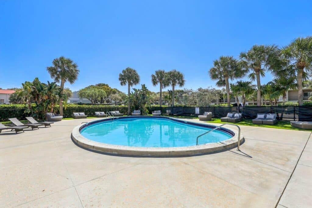 A tranquil outdoor swimming pool at Boca Raton apartments for rent, surrounded by lounge chairs and palm trees under a clear blue sky. The concrete deck is perfectly empty, creating a serene and inviting atmosphere.