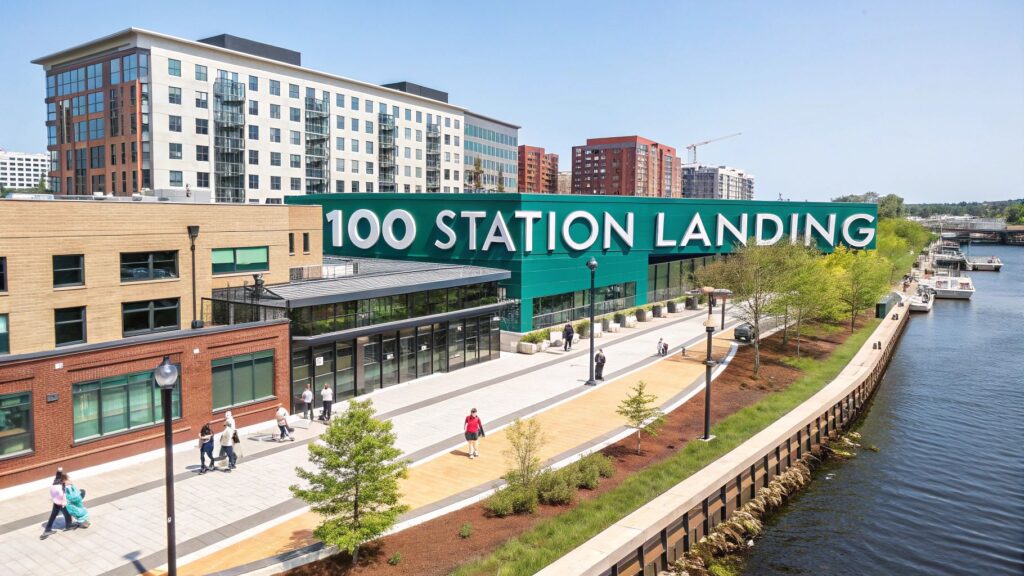 A modern riverside building with a large sign reading "100 STATION LANDING," surrounded by walkways, trees, people walking, and multi-story buildings in the background on a sunny day.