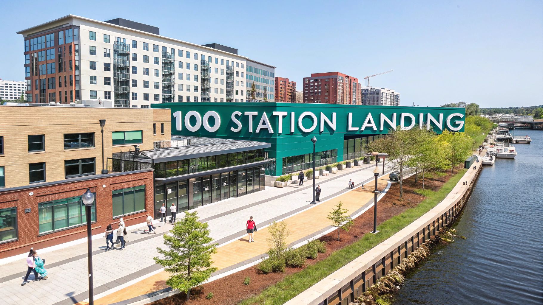 A modern riverside building with a large sign reading "100 STATION LANDING," surrounded by walkways, trees, people walking, and multi-story buildings in the background on a sunny day.