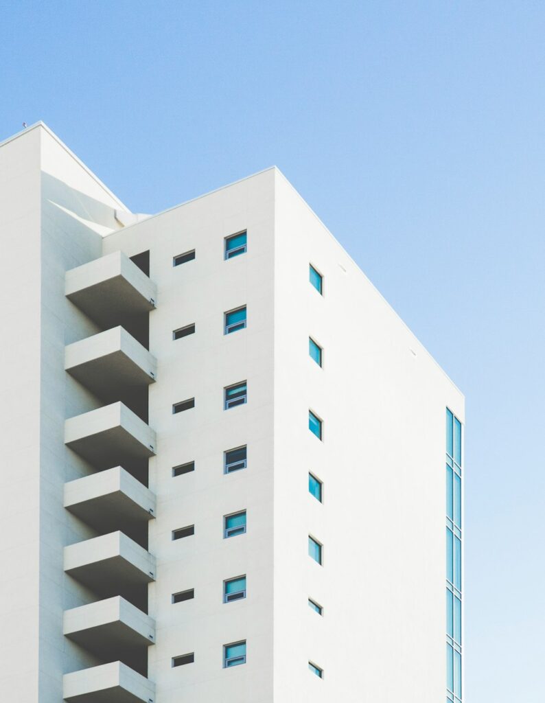 A modern white building with small square windows and protruding balconies, set against a clear blue sky. The minimalist architecture features clean lines and a smooth facade.