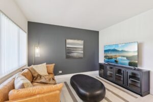 A modern living room in Apartments in Boca Raton, featuring a tan leather sofa, black oval coffee table, striped rug, dark accent wall with artwork, floor lamp, and a large TV on a black console displaying a scenic landscape near FAU.