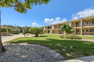 A two-story beige apartment building with balconies overlooks a green lawn, trees, and a paved walkway under a clear blue sky—perfect for those seeking Boca Raton apartments.
