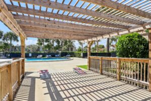 A wooden pergola casts striped shadows on a sunny patio near a swimming pool at Apartments in Boca Raton, with palm trees and lounge chairs in the background. Two cornhole boards are set up under the pergola.