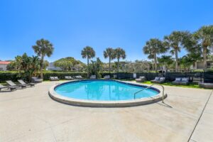 A round outdoor swimming pool, surrounded by lounge chairs and palm trees under a clear blue sky, offers relaxing seating areas and lush greenery—the perfect escape at these Boca Raton apartments.