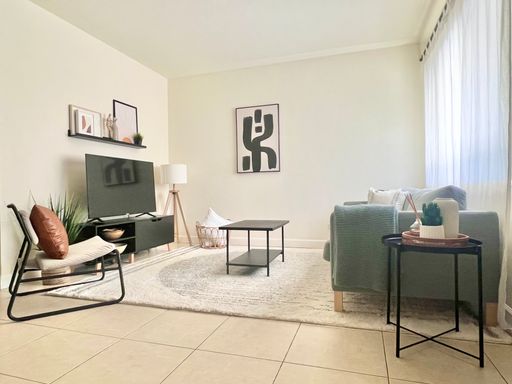Bright living room with neutral tones, featuring a green sofa, accent chair, coffee table, TV stand, abstract wall art, plants, and a large window with sheer curtains—live the good part in a space filled with natural light.