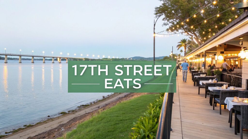 Outdoor riverside restaurant with string lights, tables set for dining, and people walking along a boardwalk at sunset; a green sign in the center reads “17TH STREET EATS.”.