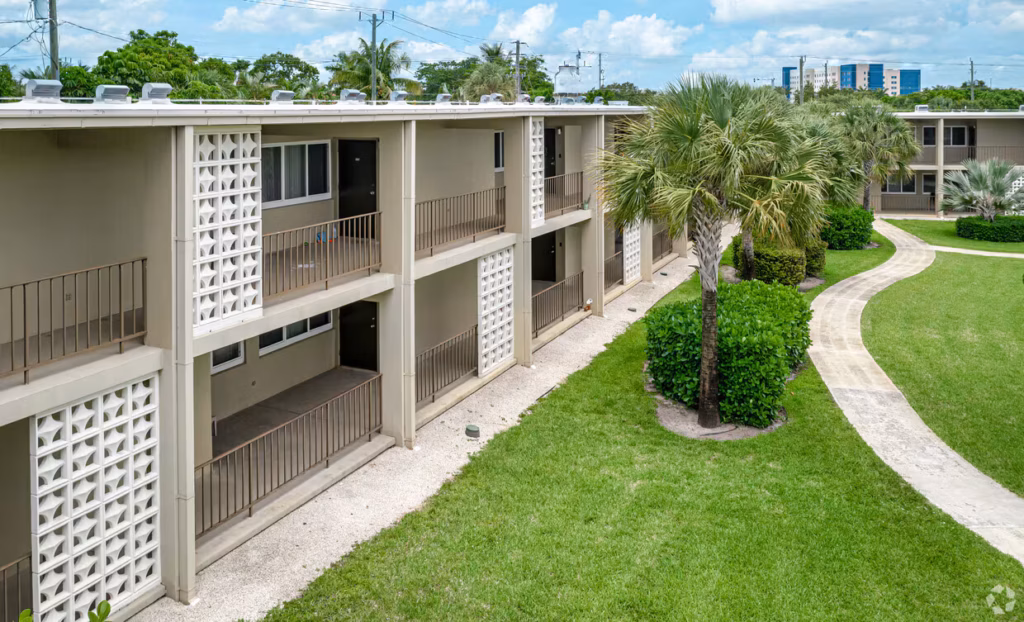 A two-story apartment building with balconies showcases modern apartment features as it overlooks a landscaped courtyard filled with palm trees, bushes, and a curved sidewalk on a sunny day.