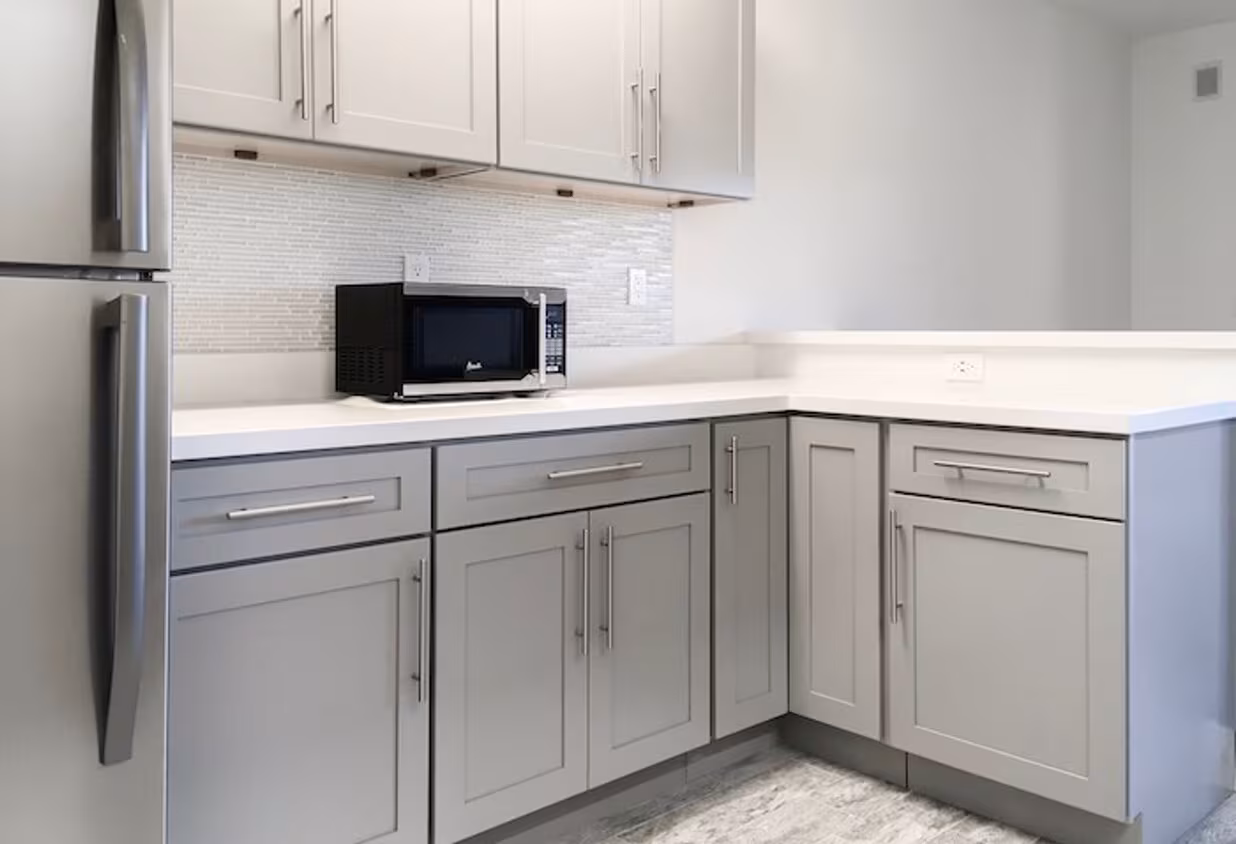 A modern kitchen highlights apartment features like light gray cabinets, a white countertop, black microwave, stainless steel handles and fridge, with a tiled mosaic backsplash that complements contemporary apartment styles.