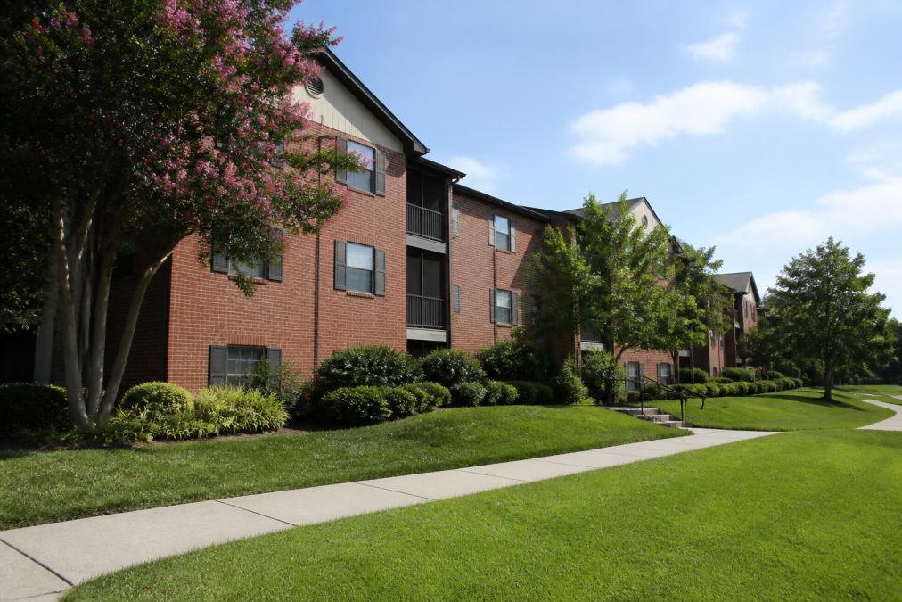 A modern brick apartment building, Kaiser Park Apartments, features large windows and balconies, surrounded by well-maintained green lawns, trees, and a curved sidewalk under a bright blue sky.
