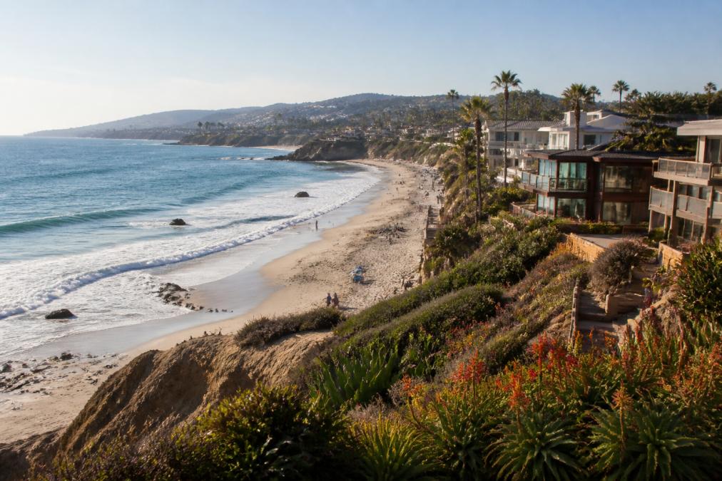 Oceanfront houses overlook a sandy beach lined with palm trees in one of the best beach towns in California. People walk along the shore as waves gently approach, while hills and more homes are visible in the background under a clear blue sky.