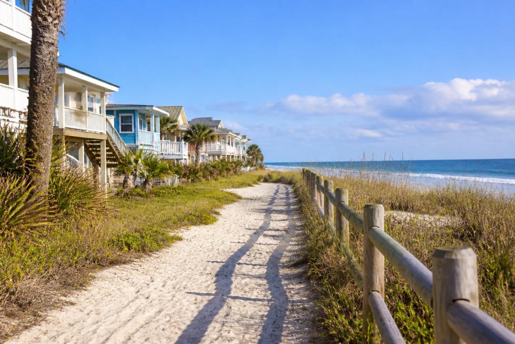 A sandy path lined with a wooden fence runs beside beach houses in one of the most affordable beach towns, leading toward the ocean, with palm trees and blue sky in the background.