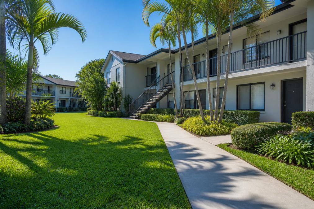 A sunny courtyard at a garden style apartment complex with green grass, palm trees, shrubs, and two-story buildings featuring balconies and outdoor staircases.