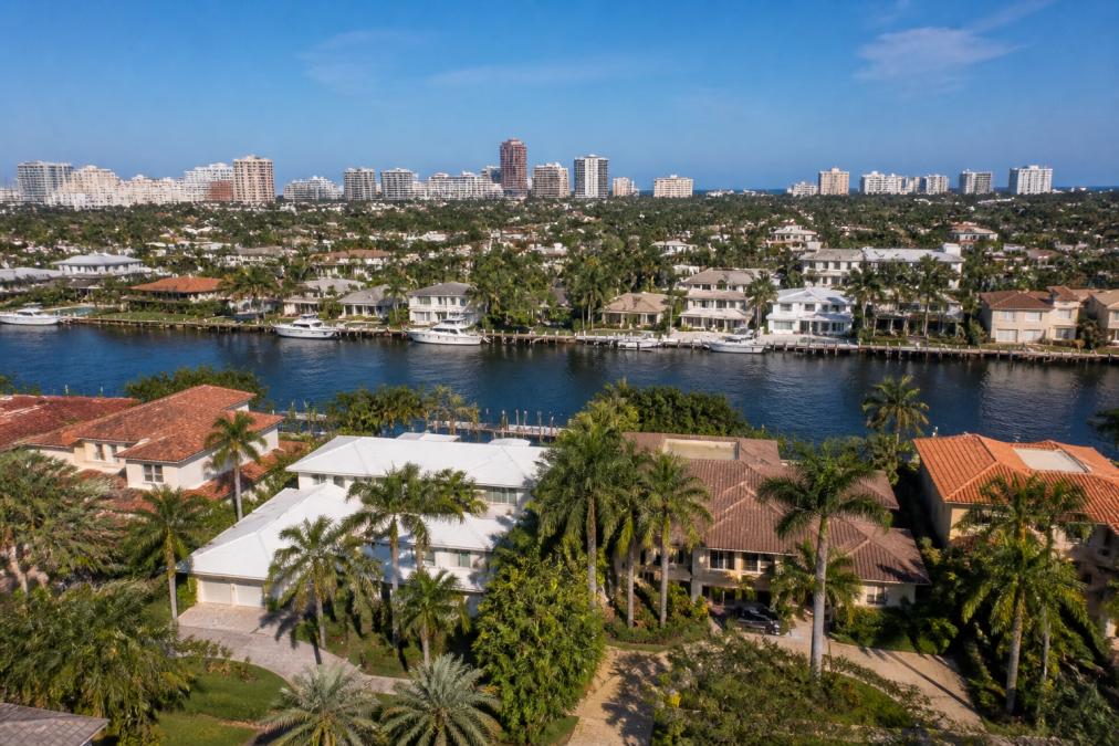 Aerial view of waterfront houses with docks and palm trees, a canal in the middle, and high-rise buildings in the background under a clear blue sky—a picturesque scene that often leads visitors to wonder: are home prices dropping in Boca Raton?.