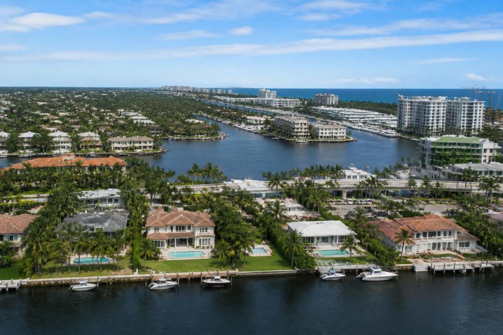Aerial view of waterfront homes with pools, palm trees, and boats docked along canals—raising the question, does Travis Kelce have a house in Boca?—with high-rise buildings and the ocean in the background under a blue, partly cloudy sky.