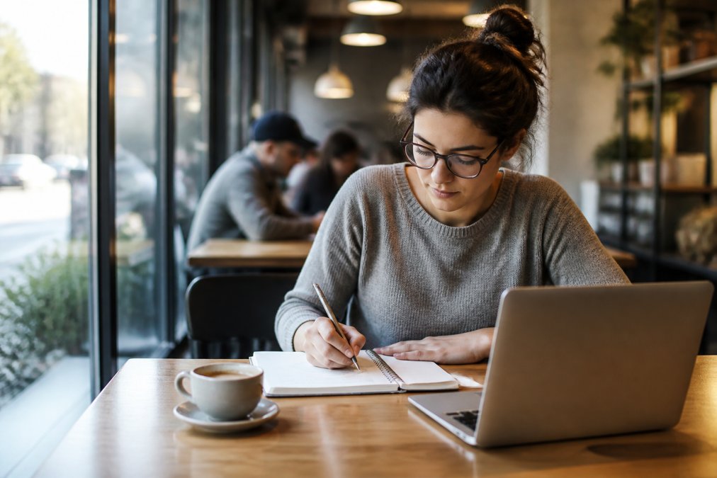 A woman with glasses writes in a notebook while sitting at a table with a laptop and coffee in a bright café, one of the popular cafes for studying.