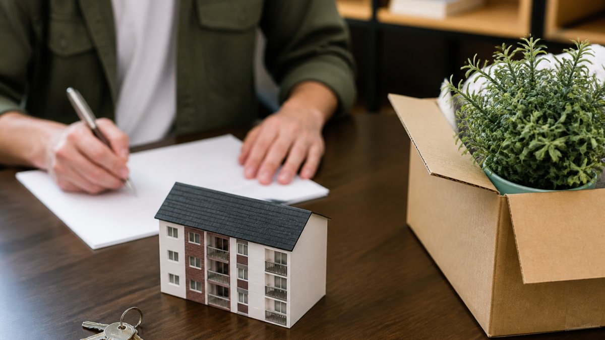 A person writes on a document regarding how to break an apartment lease in Florida next to a model building, keys, and a cardboard box containing a plant.