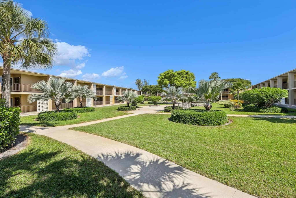 A concrete path winds through green lawns in the courtyard of these Boca Raton apartments for rent, surrounded by low-rise buildings and palm trees under a clear blue sky.