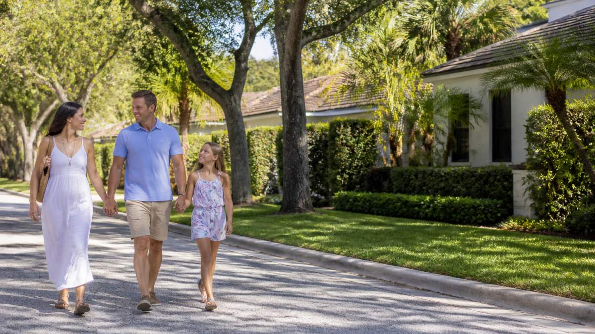 Family walking through a tree-lined residential street in Boca Raton on a sunny day, with attractive homes and well-maintained yards visible in the background