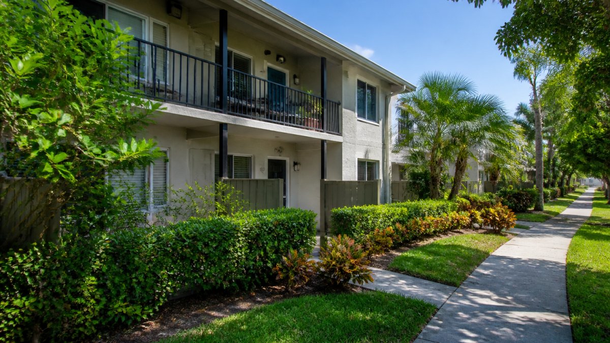 Two-story garden apartments with balconies, surrounded by landscaped hedges and a sidewalk leading into the distance.